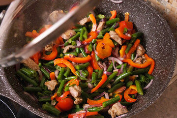 Woman lifts the lid and stirs simmering stew of green beans, red pepper, onion, and meat in a deep pan