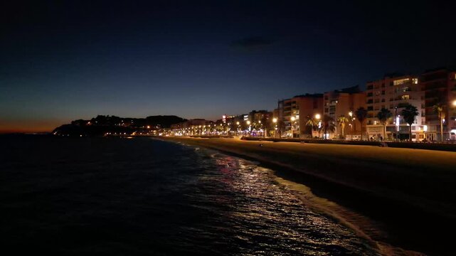 Aerial drone view of coastline town at night, moonlight reflected on sea waves, city lights, vertical background