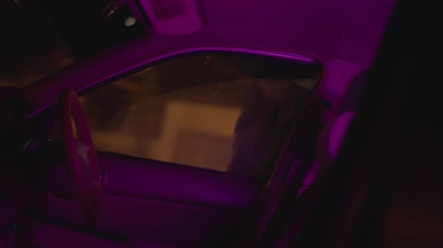 interior car at night driver adjusting posture purple neon glow bathes dashboard man in blue jacket checks mirror phone engine idles waiting tension cinematic rideshare late shift worker scene urban