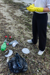 Obraz premium volunteer cleaning polluted beach, collecting plastic bottles, cups and debris in a black bag, supporting ecology, environment protection, sustainability and coastal cleanup awareness.