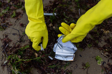 Hands in yellow gloves picking up crushed packaging and litter during beach cleanup, showing pollution, environmental damage and the importance of eco awareness and coastal protection.