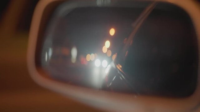 rearview mirror reflecting approaching headlights, bokeh streetlights and wet glass streaks from wiper, oncoming traffic glare and lens flare, cinematic nighttime commute from driver perspective,