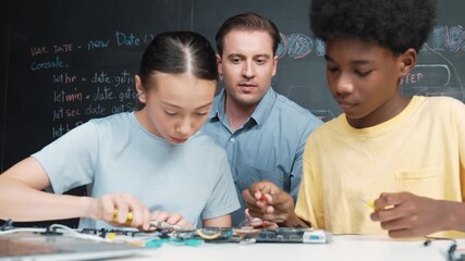 Caucasian teacher talking about electric tool while student fixing robotic model on table with electronic equipment placed. Diverse academic children learning about main board structure. Edification