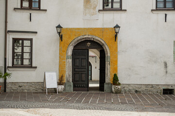 An arched stone tunnel frames a view of a white historic building with brown window shutters and a faded religious fresco. A wooden ladder hangs on the left wall of the narrow, paved passageway.