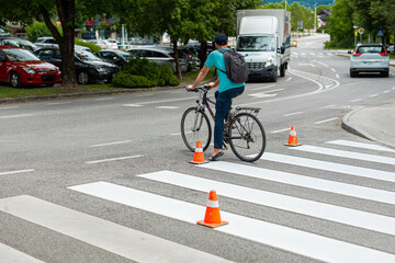 A cyclist in a turquoise shirt and backpack crosses a freshly painted zebra stripe crosswalk. Orange traffic cones mark the new white lines on the asphalt road amidst moving cars and a white van.