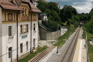 The Radovljica railway station building stands beside modern tracks and a platform. The historic white structure features decorative window surrounds and a brown gabled roof overlooking the lush