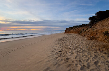 Beautiful sandy beach with red rocks and cliffs. Sunset at dusk with shallow waves on the Atlantic coast in Praia de Loule Velho, Algarve, Portugal