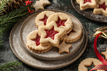 Homemade Linzer Christmas cookies filled with raspberry jam and dusted with sugar on ceramic plate with fir tree branches on wooden table