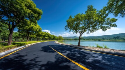 Fototapeta premium A road curves alongside a lake with trees on both sides under a bright blue sky. Mountains are visible in the background creating a natural view.
