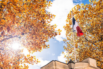 French flag waves proudly atop a stone structure, framed by vibrant autumn trees under a clear blue sky. Golden leaves contrast with tricolor stripes in a striking, patriotic scene.