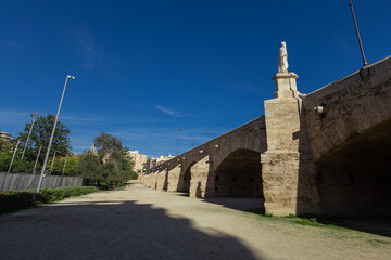 The Royal Bridge Puente del Real stretches across the Turia Garden in Valencia. This historic stone bridge features grand arches and a white statue of a saint atop a pedestal under a clear blue sky.