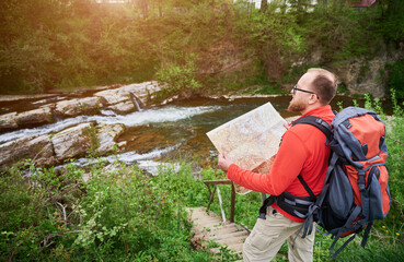 Bearded tourist man studies map while on wooden staircase by river. Traveler with grey backpack and glasses, planning outdoor adventure, hiking or climbing route.