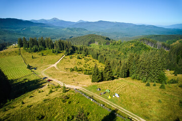 Aerial view campsite in mountains, with several colorful tents on grassy clearing. Campers relax under clear blue sky, surrounded by tall evergreens and panoramic views of distant peaks.