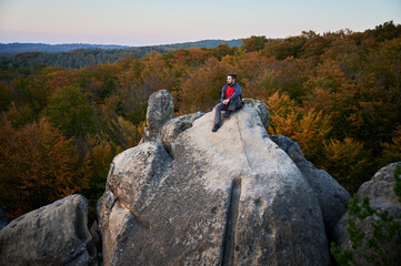Aerial view of climber sitting atop rock formation at Dovbush Rocks in Carpathian mountains, Ukraine. Sun sets, casting warm glow over autumn forest and distant hills, creating breathtaking view.