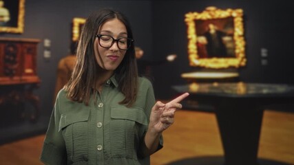 Hispanic woman points finger at painting in gallery building wearing glasses and green shirt; curiosity appreciation.