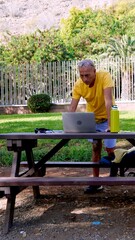 Fototapeta premium Mature man standing at a picnic table in a park, focusing on his laptop with a water bottle nearby, embodying a balanced remote work and healthy living concept