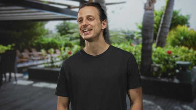 Man smiling with teeth visible and long hair pulled back, wearing a black tshirt while standing by hotel pool terrace outdoors; happiness relaxation.