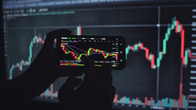 Trader analyzing online BTC price view of live trading on a smartphone with a candlestick chart, backed by a monitor displaying market data
