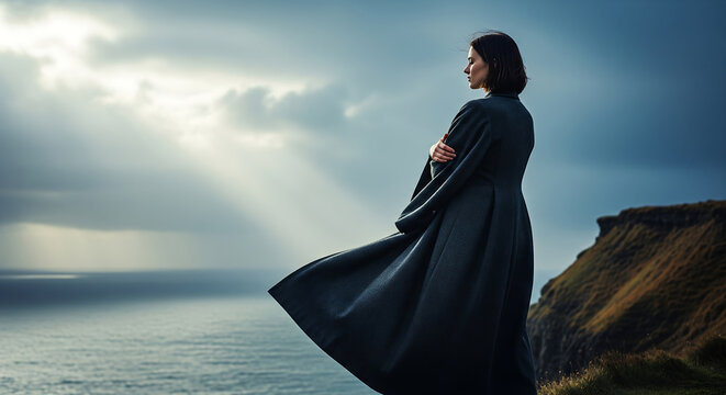 Woman in long coat standing on cliff edge looking at the ocean under dramatic sky with sun rays - Powered by Adobe