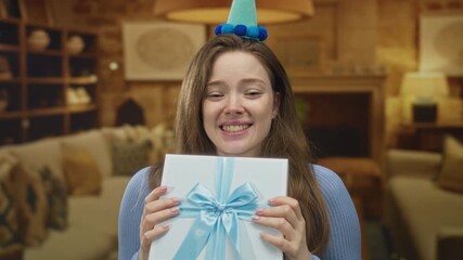 Woman holding giftbox with blue ribbon and wearing party hat while smiling in cozy living room; festive joy.