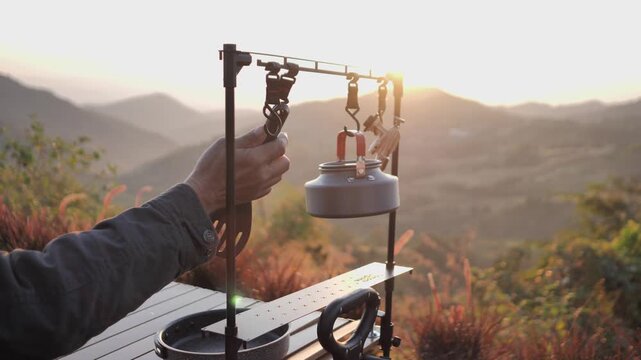A mature Asian man prepares coffee at a mountain campsite during sunrise. This outdoor lifestyle scene captures a peaceful camping morning with professional gear and a beautiful valley background.