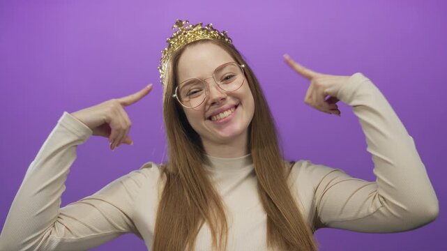 Young woman wearing crown and eyeglasses smiles and points finger to crown in purple studio; confidence.