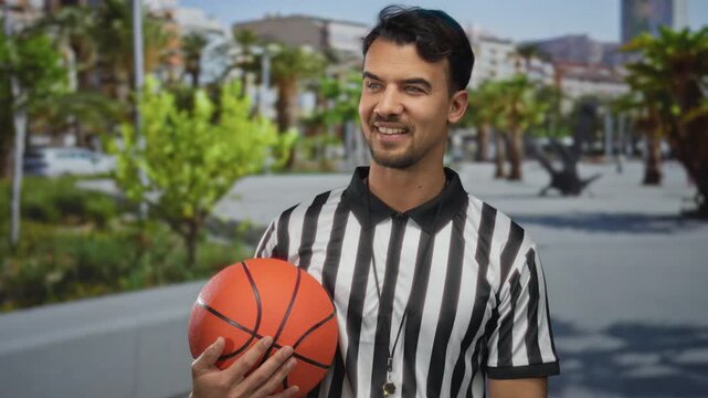 Young hispanic man outdoors holding basketball wearing referee shirt in sunny urban background with palm trees and blurred cityscape expressing sports enthusiasm.