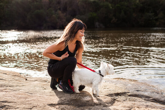 A woman kneels by the river's edge, smiling at her small white dog while enjoying nature