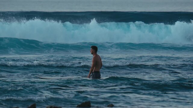 Shirtless Man with surfboard Wading Into Rolling Waves Across Shallow Reef And Rocky Shore, Relaxed Posture With Playful
