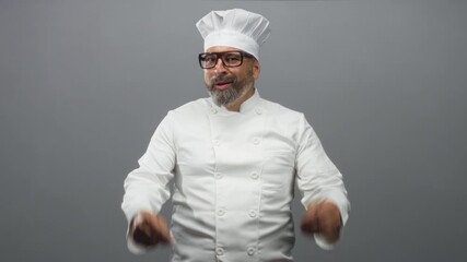 Man chef in white uniform and hat with grey hair, beard and glasses, gesturing with hands in a grey studio; confidence expertise craft.