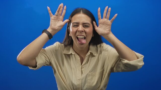 Young woman sticking tongue out with hands raised in studio with blue backdrop; playful mischief youthful energy.