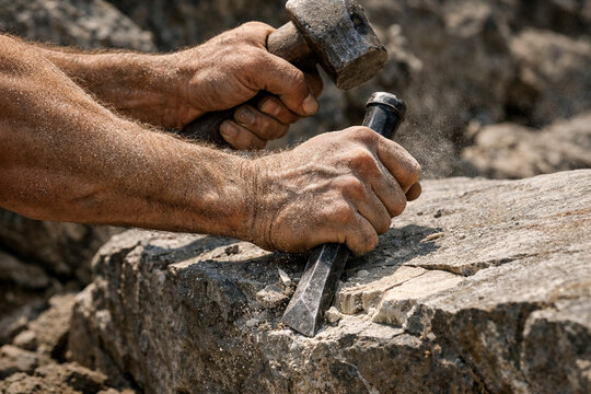 Close up of stonemason hands using a heavy hammer and sharp chisel to carve a large natural stone block.