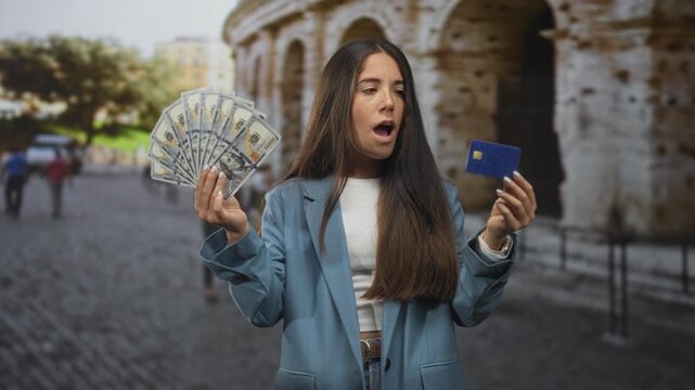 Woman holds a fan of us dollar bills in one hand and a blue creditcard in the other on a cobblestone street by a stone building; surprise temptation spending.