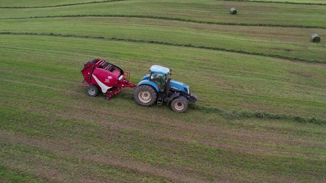 Drone side view of a blue tractor towing a red round baler along a windrow in a green meadow, with finished hay bales scattered in the distance.