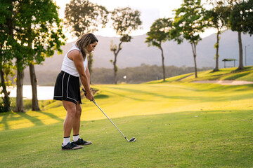 Hispanic woman focusing on her shot during a golf game on a lush green course with trees and water...