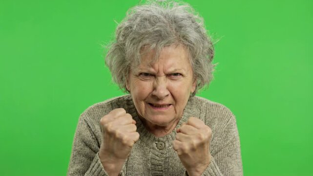 Enraged Elderly Woman With Grey Hair and Wrinkles Clenches Fists in Frustration Against A Vibrant Green Screen Background