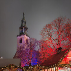 St. Mary&rsquo;s Church tower rises behind colorful illuminated trees and festive wooden stalls at the Christmas market on Alexanderplatz in Berlin on a winter night.