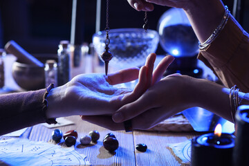 Fortune teller with pendulum reading woman's palm at table, closeup
