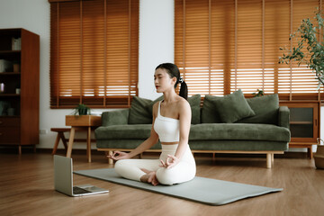 Young Asian woman meditating in lotus position at home, sitting on the floor in fitness clothing. Breathing workout to achieve relaxation, peace and mindfulness