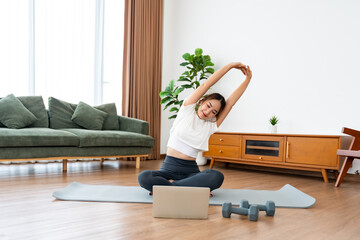Young Asian woman stretching yoga workout on exercise mat while online training class with computer laptop in living room