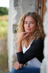 Woman with curly hair resting chin on hand by an old doorway in a rustic outdoor setting, Generative AI