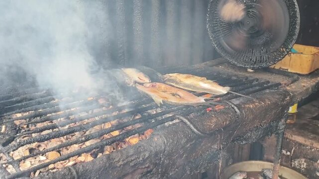 Traditional Indonesian ikan Bakar (Grilled fish) being smoked and barbecued over hot charcoal and dried coconut husks, creating a smoky, authentic flavor. Focus on cooking process.