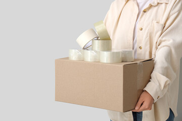 Young woman with adhesive tape rolls and cardboard box on light background