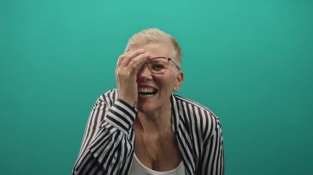 Woman laughing, hand covering one eye, wearing striped shirt and glasses in studio turquoise background; joy playfulness confidence.