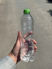person's hand holding a clear, empty plastic water bottle with a bright green cap outdoors. The bottle features a hexagonal textured pattern.