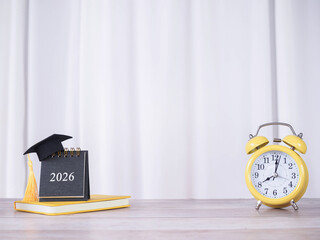 Creative desk, 2026 calendar with graduation hat on hardcover book and Yellow alarm with white curtain for background. Copy space for text, Office desk concept.