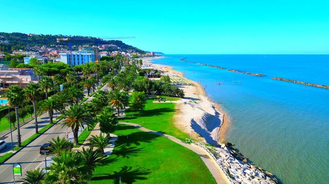 GROTTAMMARE, ITALY - SEPTEMBER 15, 2025: Aerial truck left view of Parco sul mare. Vivid green promenades and lines of palm trees meet lush rippled aquamarine Adriatic sea waters and infinite horizons