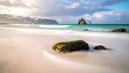 Serene coastal scene with smooth sandy beach, turquoise water, rocky outcrops, and a mountain range in the distance, under a cloudy sky
