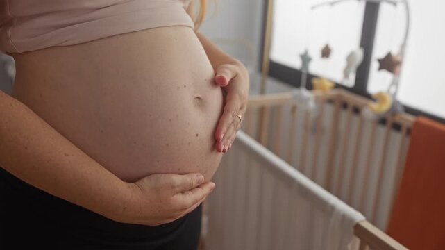 Young woman tenderly cradles bare pregnant belly with both hands in studio nursery beside wooden crib; serene anticipation.