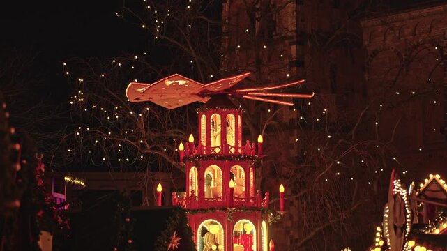 German Christmas pyramid glowing against the night sky at Bonn's M&uuml;nsterplatz. The structure features traditional craftsmanship with carved figures and a large wooden rotor at the top.
